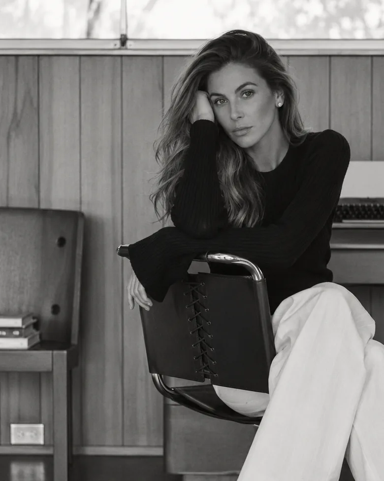 Nicola Elizabeth seated in a mid-century studio office, looking toward the camera, with a typewriter and books behind her